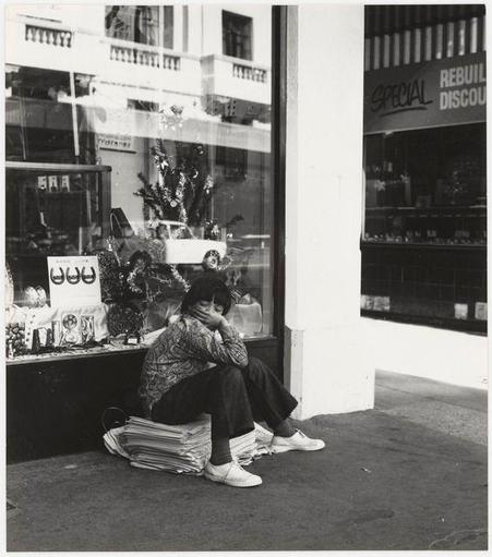A black and white photograph capturing a moment of urban life. A person sits on an improvised seat made from stacked newspapers outside a storefront that appears to be selling various decorative items, including what seems like small Christmas trees in the window display.
The individual is dressed casually with patterned clothing, dark pants, and light-colored shoes; they are hunched over slightly, resting their head on one hand which suggests fatigue or contemplation. The setting includes reflections of buildings across from this scene visible through the storefront's glass windows, indicating a busy urban environment. Above the window display hangs signage advertising "SPECIAL REBUILD DISCOUNT," suggesting that there may have been recent renovations to businesses in the vicinity.
The photograph evokes themes of solitude amidst the hustle and bustle of city life, with an intimate glimpse into personal moments set against the backdrop of commercial activity. The use of monochrome enhances the starkness of this scene, emphasizing textures like the crinkled newspapers and the individual's fabric patterns.