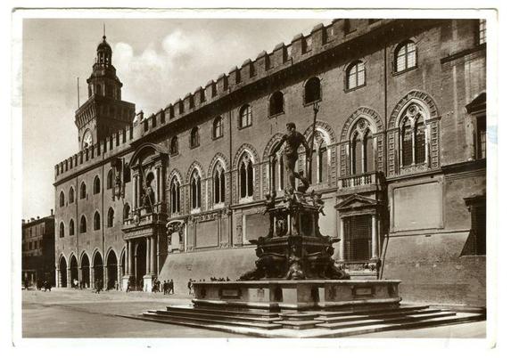 This black and white historical photograph shows a grand neoclassical building with multiple arched windows, ornate cornices, and an imposing clock tower. In the foreground is a bronze fountain featuring statues on top, surrounded by several stone steps leading down to what appears to be a public square or plaza. A few individuals can be seen walking around in the distance near the base of the structure. The sky above has a cloudy appearance, suggesting overcast weather conditions at the time this photograph was taken.