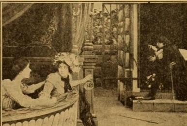 A surviving film still. The scene appears to be set outdoors, possibly a garden or courtyard, with a balcony or viewing area in the foreground.
In the foreground, two women are positioned on what appears to be a curved balcony or a decorative structure. The woman on the right, wearing a hat adorned with flowers, is looking towards the right side of the image with a surprised or concerned expression. She is partially leaning forward, her arm extended towards a decorative element on the structure. The woman on the left is positioned behind her and to the left, slightly out of focus. She's also looking towards the right, her expression less defined.