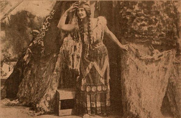 Anna Rosemond as Pocahontas.
The image is a sepia-toned photograph depicting a woman in what appears to be a Native American-inspired costume. The woman is the central figure, positioned slightly off-center to the right. She has long, braided hair, and is wearing a headband, a fringed top, and a skirt adorned with decorative elements, possibly metal discs. Her arms are outstretched, one hand raised to her head, the other resting on the rough, textured surface to her right.
The background is composed of a backdrop that mimics natural elements. To the left, there's a large, textured structure resembling a rock formation, with a section of fabric patterned with a leopard-like print draped over it. Partially obscured within this formation, a smaller figure is visible. To the right of the woman, a similar textured surface provides a backdrop. A small, simple wooden box is placed at the woman's feet.