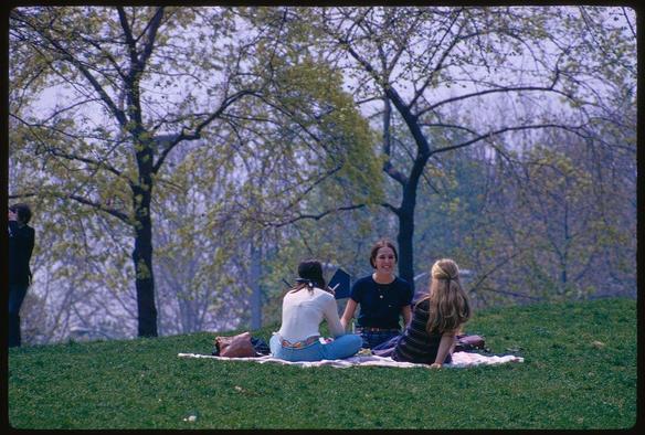 The photograph captures a serene moment within Central Park during springtime in May 1970, showcasing the lush greenery and vibrant atmosphere of New York City's iconic park. In this picturesque scene, individuals can be seen leisurely enjoying their time outdoors on blankets laid out on the grass.

Three people are prominently featured: one sitting with crossed legs wearing a white top paired with blue jeans adorned with colorful patches; another standing in casual attire donning a dark sweater and jeans; while the third person is also seated but dressed differently. The setting includes various trees, some of which have budding leaves hinting at new growth during spring.

The image encapsulates the essence of Central Park's charm as captured by renowned photographer Toni Frissell through her lens in 1970.