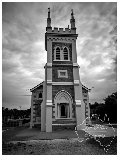 A black and white, low angle photograph of the historic Manaranga Church, featuring a prominent central tower with Gothic style pointed arch windows and battlements at the top, capped by two small spires. The church facade is built from contrasting materials, possibly stone and render. The sky is heavily clouded, adding a dramatic, moody atmosphere.