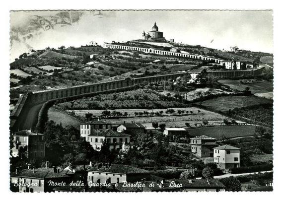 The image depicts a historical black and white photograph of the landscape surrounding Monte della Guardia in Bologna, Italy. The focal point is an imposing domed structure at the top right corner, which appears to be Basilica di S. Luca (St. Lucy's Basilica). Surrounding it are various buildings that seem like part of its complex or related structures.

The terrain slopes gently upward and away from this central building towards a long series of arches forming what looks like an aqueduct running along the middle left side, leading down to cultivated fields with terraced rows indicative of agricultural use. Below these fields is another cluster of buildings resembling residential housing.

In front of them lies more open land that could be vineyards or orchards, given their orderly arrangement and spacing between plots. The entire scene suggests a rural setting possibly from the early 20th century judging by its style, as it's labeled with "Monte della Guardia e Basilica di S. Luca" in Italian.

The image has been framed to look like an old photograph or postcard, complete with signs of age such as creases and discoloration along the borders which add to its vintage appeal.