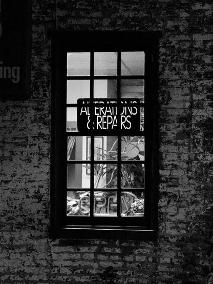 A black-and-white nighttime photo of a lighted window with a sign saying “Alterations & Repairs.” the window is in a brick wall with weathered paint.