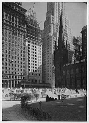 This black and white photograph depicts a scene from New York City, specifically Trinity Churchyard in 1932. The image shows the iconic skyline of Manhattan with prominent buildings such as the Empire State Building visible in the background.
In the foreground, there's an expansive graveyard filled with numerous gravestones that create a patterned landscape across the area. Some tombstones are closely packed together while others have more space between them. A few trees and shrubs can be seen scattered throughout the cemetery grounds, adding to the overall scenery. The sky above is clear, indicating good weather conditions during the time of capture.
The photo captures the contrast between the historical graveyard setting and the modern skyscrapers that define New York City's skyline. This image offers a glimpse into both the past and present urban landscape of Manhattan.