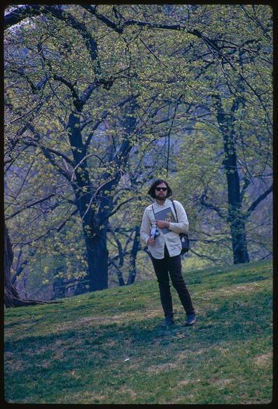 a man wearing sunglasses standing on a hill in front of trees holding several objects including two cameras and books one book titled "new york" with his right hand. he is carrying the items while looking down at them and appears to be walking through a park during springtime as indicated by the flowers blooming all around him