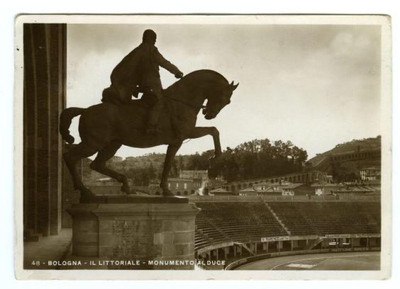 The image is a sepia-toned photograph showing an equestrian statue, with the figure of a man seated on horseback captured in mid-motion as if jumping. The rider appears to be dressed historically or formally, possibly indicative of a significant historical personage commemorated by this monument.

In the background, there's a stadium seating area known as "Il Littoriale," which is identified at the bottom right corner with white text on black: "BOLOGNA - IL LITTORIALE - MONUMENTO AL DUCE." This suggests that the statue might be located in Bologna and could commemorate Benito Mussolini, often referred to by his nickname 'Duce.'

The stadium appears empty or not crowded at this moment. The surrounding landscape includes buildings on a hillside and an open sky above. There is no visible audience within the stands.

This image carries historical significance as it captures part of Bologna's heritage during Italy’s Fascist period, with "Il Littoriale" being one of Mussolini’s personal projects for promoting Italian nationalism in cities along the coastlines that were to become important ports under a unified Italy. The statue could represent an aspect or achievement associated with his regime.

Please note that mentioning this image within any context should be done sensitively, considering its historical and cultural weight.