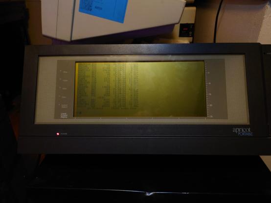 A picture of the Apricot portable sat on top of a cabinet, showing the old greyscale wide aspect LCD showing the DOS readout of DIR on the boot disk, showing all the top level files and directories.