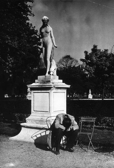 The image depicts a black and white photograph of an outdoor scene featuring a large statue on a pedestal, likely in a park or garden setting. The statue is of a female figure with classical attributes, possibly representing the goddess Venus, standing elegantly above her smaller companion, which appears to be Cupid (Eros). Both figures are nude and stand out starkly against their surroundings.
In contrast to the grandeur of the statues, there's an individual seated on a chair beside them. This person is bent over in distress or deep thought, with head bowed down towards what seems like luggage next to him, suggesting he might be traveling or waiting for someone. The surrounding area appears well-maintained, with manicured bushes and trees providing a serene backdrop.
The overall mood of the photograph juxtaposes classical artistry against human vulnerability and solitude, creating an evocative scene that invites contemplation on various themes such as beauty versus suffering, permanence in contrast to transient existence, or perhaps even the dichotomy between public displays of grace and private moments of despair.