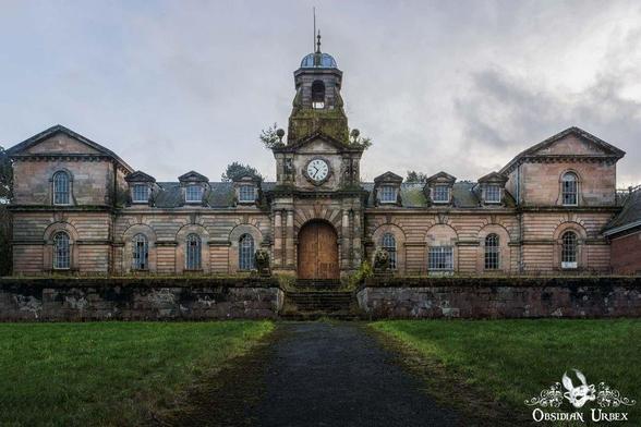 A large, abandoned brick building with a central clock tower, arched doorway, and overgrown vegetation on the roof and steps, set against a cloudy sky.