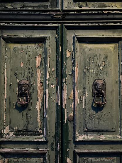 Closeup photo of the central panels of a set of panelled wooden double doors. They are painted a dark mossy green, but the paint is largely flaking and fading, revealing flashes of dark blue paint and rough blond wood underneath, and they look like no one has opened them, let alone cared for them, for decades or centuries. There are matching black iron door knockers on both halves of the door, in the shape of stern faces, with the knocker-handle-bits hanging down and casting u-shaped shadows. There is a feeling of dereliction and total neglect, and I think some people might find it a bit spooky-looking and Gothic horror-y, but I think it looks intriguing and mysterious and like an invitation to heart-pounding spectacular adventure, and I would very much like to go inside.