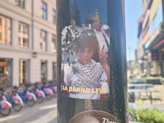 A photographic sticker on a black pole shows a young boy with a keffiyeh around his neck, holding a branch and giving a peace sign. Yellow text at the bottom reads "LA BARNA LEVE" (Let the children live).