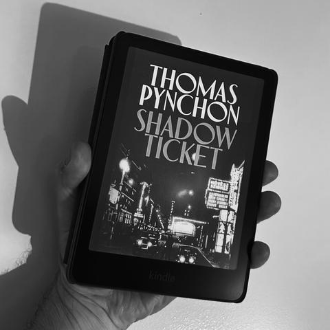 A hand holding a Kindle displaying the book cover of "Shadow Ticket" by Thomas Pynchon, set against a black and white background. The cover features a nighttime city scene with illuminated signs.