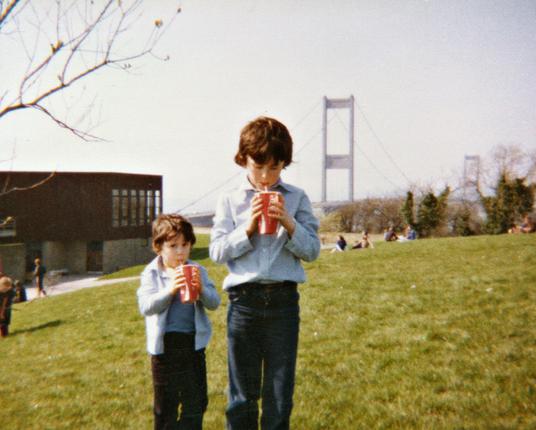 A scan of a colour photograph from around 1980, a little faded with time. We're on a grassy bank that gently slopes down to the left. In the foreground are two young boys aged roughly 5 and 10, both with dark trousers, both with light blue tops although the older boy is wearing a shirt and the younger has a t-shirt with thin jacket over the top. Both boys are also holding large Coca Cola-branded disposable cups, gripping them with both hands and sipping on straws. They've clearly just been to a service station and part of the back of a building that may belong to it is on the left at the bottom of the shallow slope of grass. There are a few people out sitting on the grass so it's a warm enough and pleasant enough day for it although any blue in the sky has been washed out to white with time. Over the boys' shoulders to the right in the distance there are the two upright concrete towers that support a suspension bridge that crosses a river out of view.