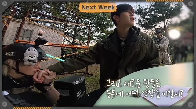 Seungmin looks out at the battlefield while soldier Bang Chan crouches beneath him, holding his wrist and kissing his hand.