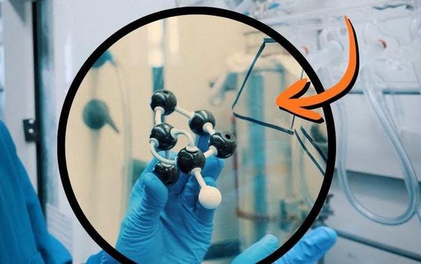 Scientist in blue gloves holding a molecular model structure with black and white spheres representing atoms connected by rods, demonstrating chemical bonding in a laboratory setting.