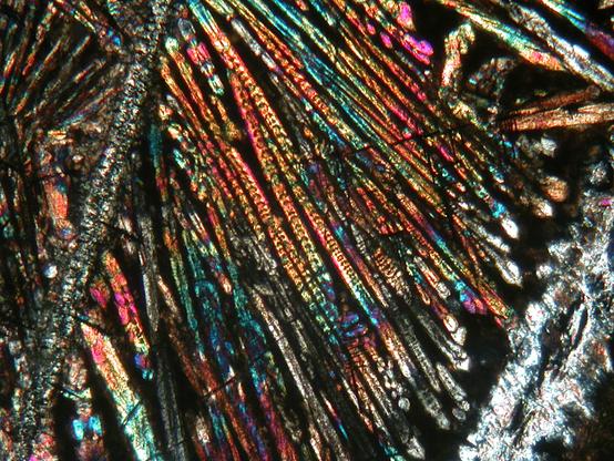 A view of a rock thin section taken down a polarizing microscope using crossed polars. The view consists mostly a long, feathery, rainbow-coloured crystals (clinopyroxene) in a matrix of fine-grained dark brown material, with two larger, long linear crystals of greyer mineral (serpentinized olivine) running NNE-SSW at the left and right sides of the view.
