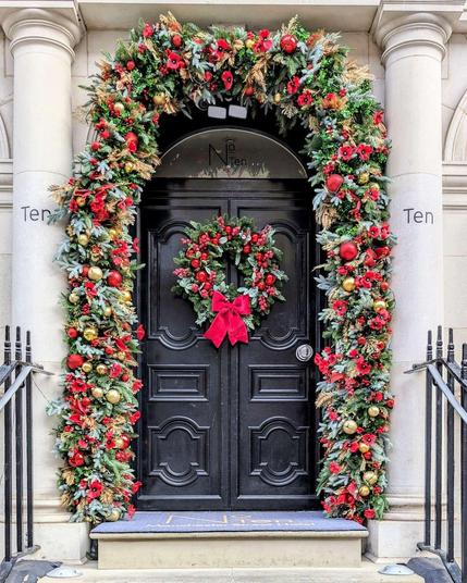Vertical close-up of a black London Christmas door ("No. 10") framed by a lush garland of red poppies and gold ornaments, featuring a matching wreath with a red velvet bow.