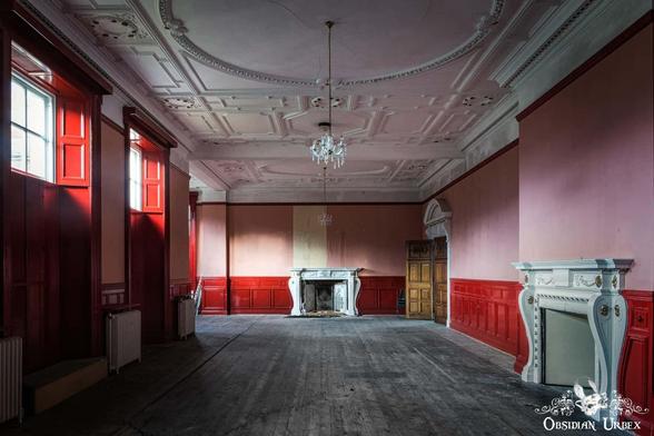 An empty, decaying room with red-paneled walls, a fireplace, wooden floor, chandelier, and tall windows letting in natural light.
