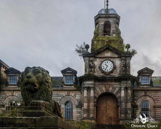 A weathered stone lion statue sits in front of an old, abandoned building with a central clock tower and overgrown vegetation.