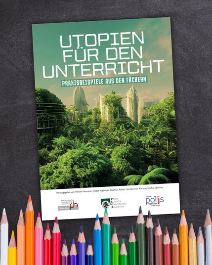 Titelbild einer Broschüre mit der Aufschrift „Utopien für den Unterricht – Praxisbeispiele aus den Fächern“. Zu sehen ist eine futuristische Stadt mit hohen, begrünten Gebäuden inmitten dichter, grüner Vegetation. Die Broschüre liegt schräg auf einer dunklen Tafeloberfläche, im Vordergrund unten sind viele bunte Buntstifte angeordnet.