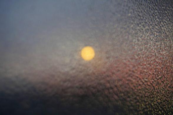 The photograph shows a hazy light that appears as a bright, blurred spot. The background is rendered in delicate shades of purple and blue, suggesting that this may be a scene at sunrise or sunset. The blurring effect gives the impression that the image could have been taken through wet or frozen glass, giving it a soft, dreamlike quality.