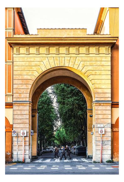 The photo is of city road framed by an archway, with pedestrians crossing it. The arch is painted in shades of yellow and orange, which frames the street. It is set against buildings with a similar colour palette.
Inside, through the arch, a street lined with tall, green trees is visible, creating a tunnel-like effect with the tree leaves. Three people are crossing the street at a crosswalk, appearing as small figures against the backdrop of parked cars and green trees.