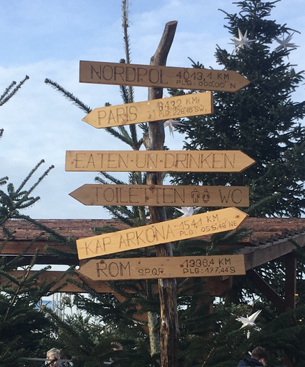 Holzwegweiser auf dem Weihnachtsmarkt in Wismar. Oben Nordpol - 4013 km, Paris 843 km, ein Wegweiser zur „Freßbude“ Eaten un Drinken, links zu den Toiletten, unten nach Kap Arkona 154 km.