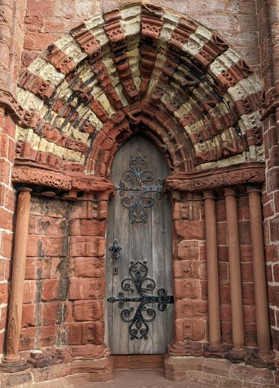 A narrow grey oak door composed of five vertical boards is embellished with enormous, elaborate iron hinges. A deep pointed arch frames the door. Built from alternating red and white carved sandstone blocks, the arch is wider than the door and gloriously gaudy. It is supported by three slim sandstone pillars on the right side. On the left there is a single pillar, plus a gap where at least two pillars used to stand.