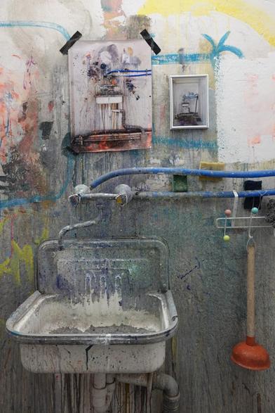 A photograph of a washbasin in an atelier with two fotos of the basin hanging there instead of a mirror. The basin is for washing-of brushes, so its full of colours.