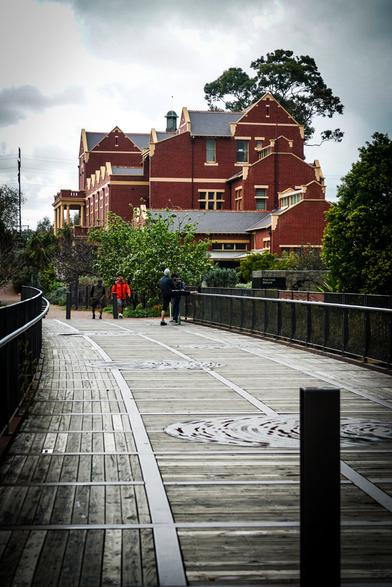 A long wooden pedestrian bridge leads toward a large, multi-story red brick building. The bridge is lined with black metal railings and has dark, circular inlays in the wood planks. Several people are walking on the bridge, receding into the distance. The brick building features multiple windows, gabled roofs, and appears to be surrounded by trees and foliage. The sky is overcast and grey, suggesting a cloudy day. Provided by @altbot, generated privately and locally using Gemma3:27b