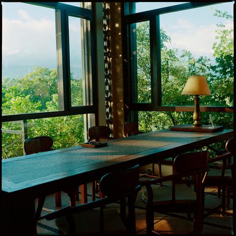 A long wooden table with chairs sits beneath large windows overlooking lush green trees and distant hills. Sunlight streams in, casting shadows across the table. A lamp with a beige shade rests on one end. The room has a serene, natural ambiance with traditional wooden furnishings.