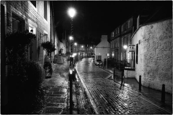 Eine atmosphärische Schwarz-Weiss-Aufnahme einer historischen Kopfsteinpflastergasse bei Nacht. Die nassen Oberflächen reflektieren das Licht der Strassenlaternen, was zu starken Kontrasten und einer tiefen Bildwirkung führt.