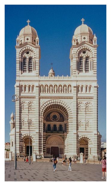 Color photograph of the Cathédrale Sainte-Marie-Majeure de Marseille. In front of the neo-Romanesque-Byzantine church, several people are standing, either on their way into the church or taking photographs of it. The church façade, made of alternating light and dark stone, is surrounded by a deep blue sky.