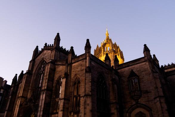 A colour photograph in landscape format of St Giles' Cathedral.

The picture is taken looking up at a steep angle. The crown shaped spire is illuminated and is in the middle of the picutre to the right. The rest of the structure is darker, lit only with available light. There is a large stained glass widow dominating the image to the left, and several masonry pinnacles can be seen along the edges of the structure.

The sky behind the cathedral is a light purple in the dusk.