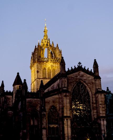 A colour photograph in portrait format of St Giles' Cathedral.

The picture is taken looking up at a steep angle. The crown shaped spire is brightlyilluminated and is in the middle of the picutre to the left. The rest of the structure is darker. There is a large stained glass widow dominating the image to the right, and some panels within the window are shining with light behind, inside the cathedral. There are several masonry pinnacles along the edges of the structure.

The sky behind the cathedral is a purple-blue.