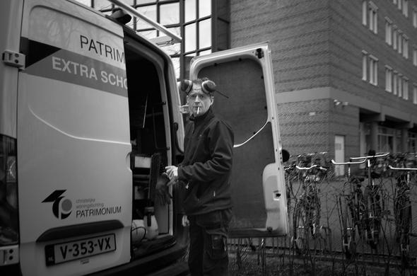 A man stands beside an open van labeled "PATRIMONIUM" while holding a cigarette. He is wearing work clothing and gloves. In the background, bicycles are parked against a fence next to a building. The image is in black and white