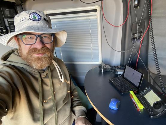 A bearded man wearing a floppy hat and glasses sits next to a table with a ic7100 radio, a Morse key, and a tiny computer