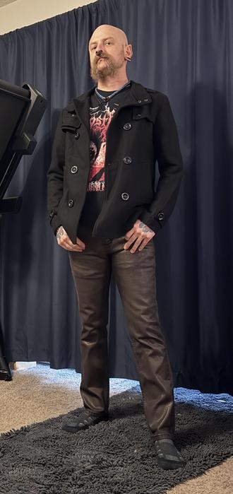 A person with a shaved head and a beard stands confidently in front of a dark curtain. They wear a black double-breasted jacket over a graphic t-shirt, paired with brown pants. Visible tattoos are on their hands, and they have multiple facial piercings.