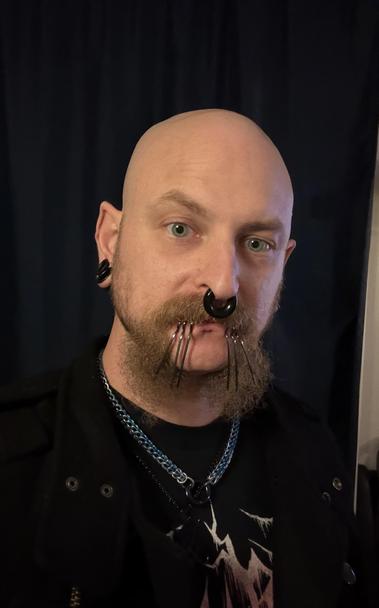 A bald individual with a distinctive style features multiple facial piercings, including a septum ring and metal labret spikes. They are wearing a black jacket over a graphic t-shirt, and a chain necklace. The background is dark.