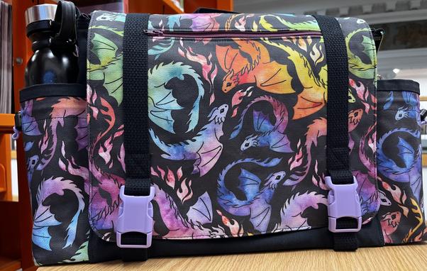 A black messenger bag with two side pockets, printed with delightful rainbow-watercolor flying dragons.  The bag has two purple buckles and a purple zipper in the flap.