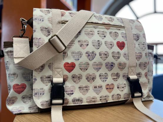 A cream messenger bag with two side pockets, printed with repeating heart shapes made of library due-date cards in various tones of cream/pink/red.