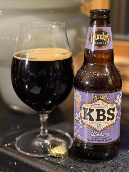 A snifter glass full of dark beer is next to a brown bottle with a light purple neck and body label. The letters KBS are front and  enter in a tan field