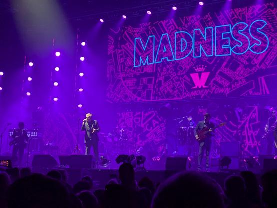 Suggs belting out on stage with a big Madness logo