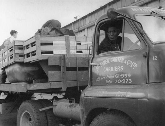 The black and white photograph depicts a vintage delivery truck from McEwen, Carter & Co. Ltd., which is positioned at the back of a wooden trailer loaded with large items such as barrels or containers. The driver's side door of the vehicle is open, revealing a person sitting inside. On top of the trailer, individuals are seen handling and maneuvering what appears to be crates or equipment.

The truck has visible contact information for McEwen Carter & Co., Ltd Carriers on its backside including phone numbers: "Phones Petone 61-959 Wgton. 70-973," which indicates the company's locations and operational centers in different regions. A smaller inscription reads, "H.M.C.I.O.T.S." beneath the main information.

The setting is an open area with a wooden fence or wall structure behind the truck, giving it an industrial feel typical of cargo handling areas at docks or warehouses during that era.

Additional context about this image can be found in Wellington wharves as mentioned in the source link provided.