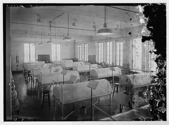 The image depicts a black and white photograph of an interior space that appears to be a classroom or laboratory. The room is filled with rows of desks, each equipped with what seem to be drafting tables or workstations, as evidenced by the angled surfaces on which papers are placed. Above these stations hang pendant lights from the ceiling.

The architecture suggests it could be part of an older institutional building, featuring large windows allowing natural light to enter and a high ceiling. The walls have several pipes running along them, adding to its utilitarian appearance. In the foreground, there is what seems like medical or scientific equipment on carts that can move around.

The room has a somewhat bare look with minimal decoration; however, it appears well-used for educational purposes as evidenced by papers and notes scattered across the surfaces of desks.

Additional information about this image suggests it could be from American University in Beirut between 1898 and 1946. It is part of an archive or collection related to medical education at that institution during those years, specifically associated with individuals named Eric and Edith Matson who were likely significant figures within the context of these photographs.

This historical setting provides insight into educational practices and facilities in a specific time frame and region, highlighting advancements and changes over decades.