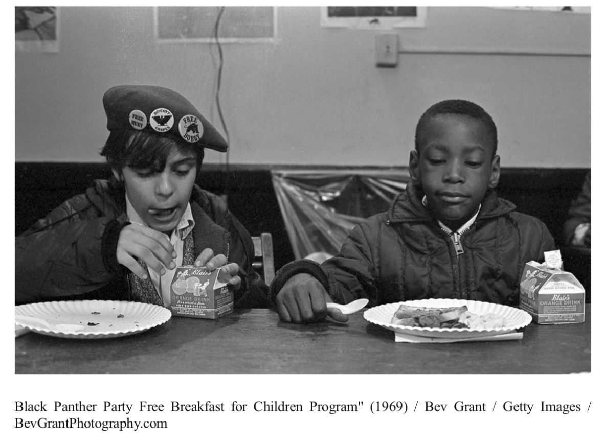 1969 photohraph of two young boys, one black and one white, eating breakfast together in a cafeteria. Credit: “Black Panther Party Free Breakfast for Children Program" (1969) / Bev Grant / Getty Images / BevGrantPhotography.com