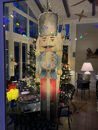 A colorful window vinyl transfer of a large nutcracker has been applied to a sunroom slider.  It has a blue coat and red pants, a very handsome mustache and scary square teeth.  Behind it a decorated and lighted tree is visible, and outdoor colorful lights also reflect on the glass.