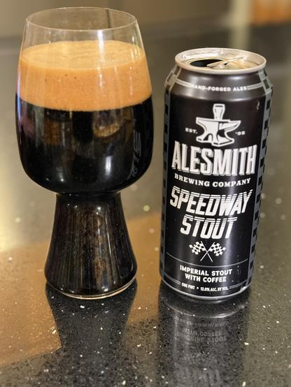 A tall stout glass full of dark beer with a thick dark tan head. The black can has white lettering, an anvil and crossed raceway finish flags