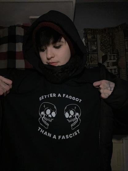 A person is wearing a black hoodie with a graphic featuring two skulls and the text "BETTER A FAGGOT THAN A FASCIST." The individual has short dark hair, wearing a beanie, and appears to be in a dimly lit Rv